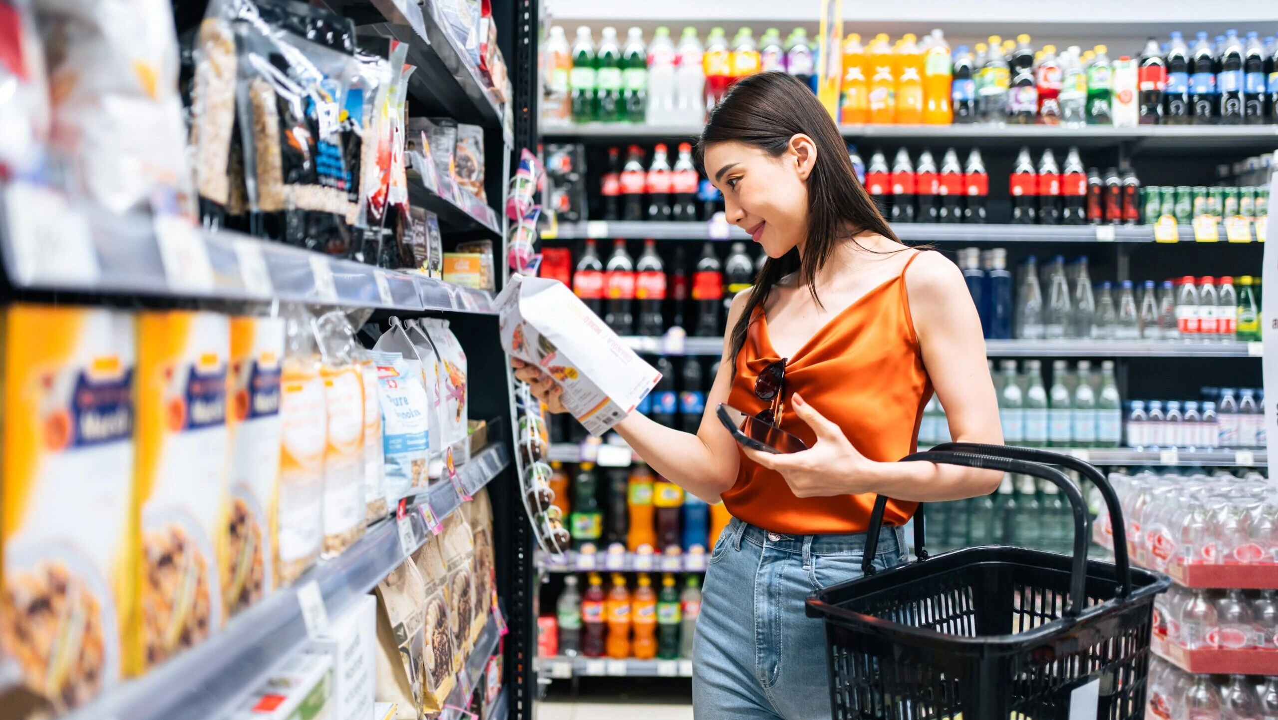 Customer shopping in a grocery store aisle, reading a food product label while holding a shopping basket, with shelves stocked with beverages and packaged goods.